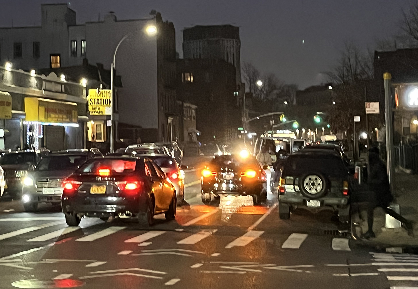 A busy street at night with a painted bike lane between the traffic lane and curbside parking. The bike lane is blocked by double parked cars and trucks.