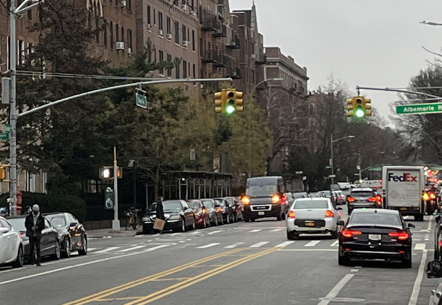 Looking down a two-way street with a bike lane on each side, looking towards an intersection. Double-parked cars and delivery trucks block the bike lane in several spots on both sides.