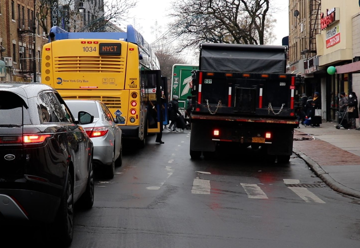 A bus, stopped in the travel lane, letting off passengers, next to a dump truck that's blocking the bus stop. Further ahead, another parked truck halfway blocks the travel lane, and car traffic is backing up behind the bus.