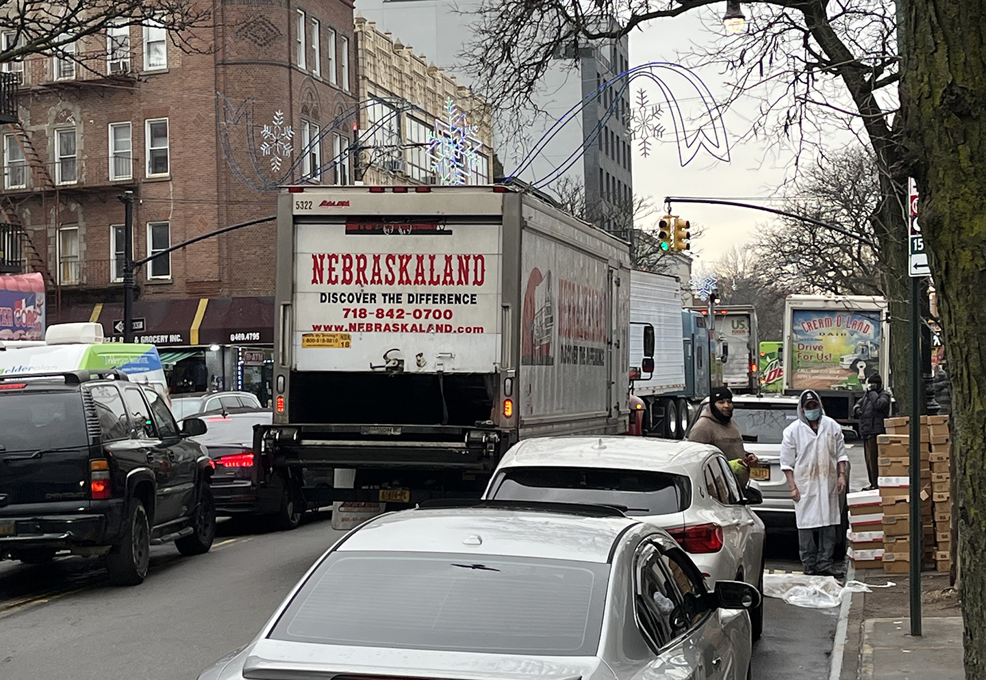Men unload groceries from a double-parked truck, while cars squeeze around on the wrong side of the street. Futher down the street, only trucks are visible, including a tractor-trailer that appears to be having trouble getting through.