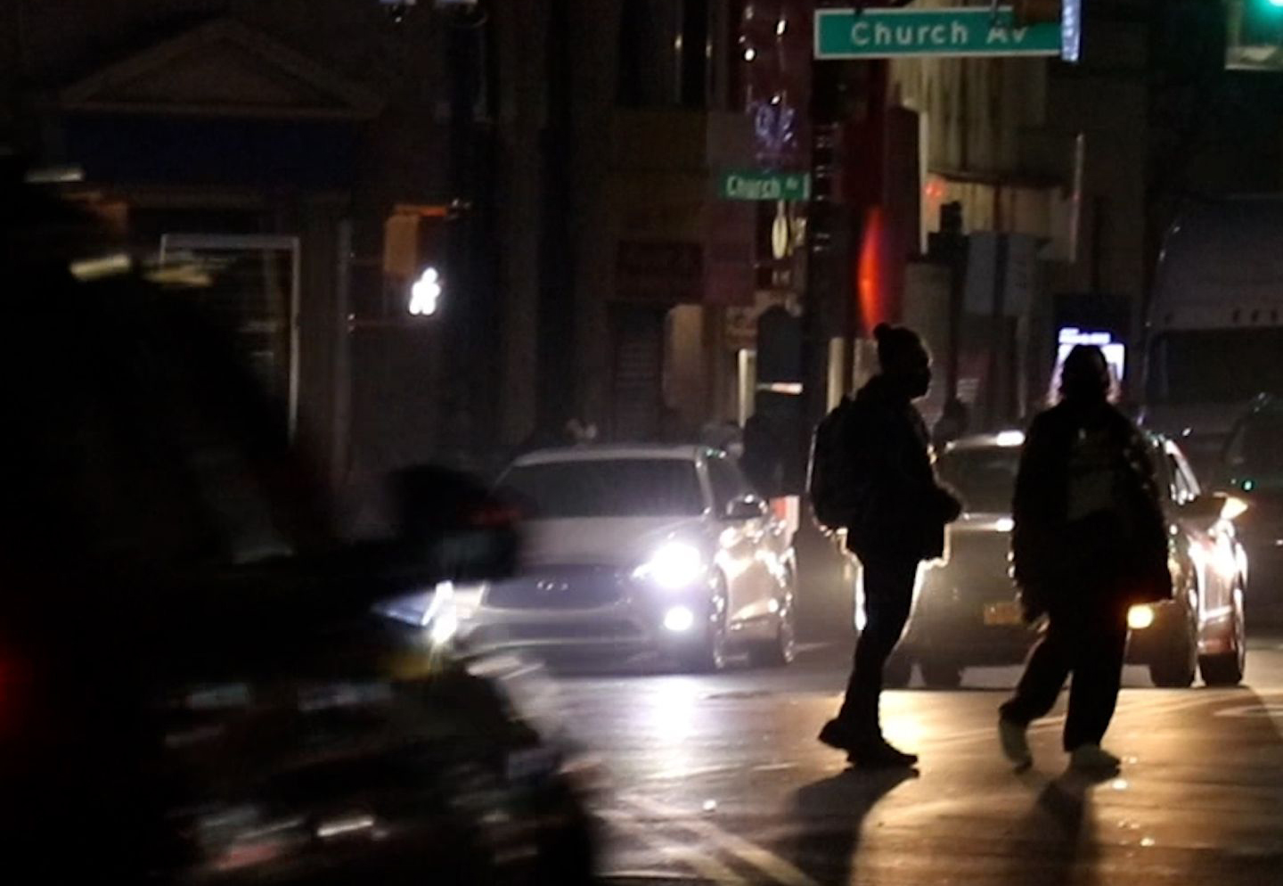 Two pedestrians, crossing an intersection in the dark, silhouetted by the headlights of cars in the background.